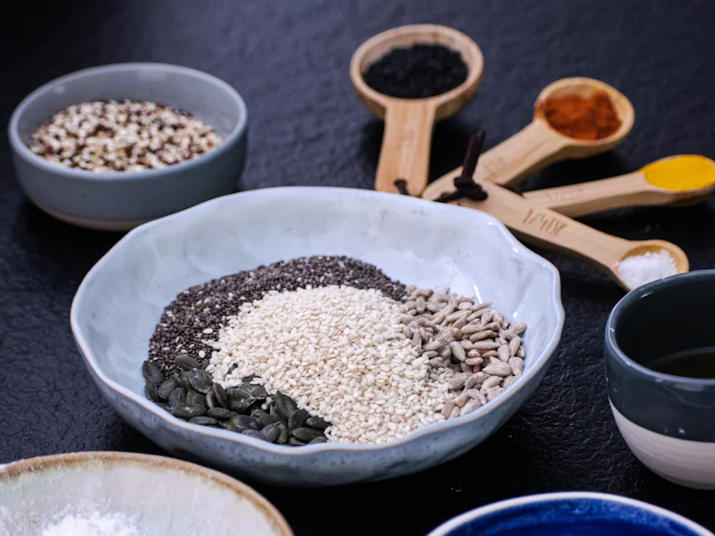 Bowls of seeds, grains and ground spices arranged on a dark surface.